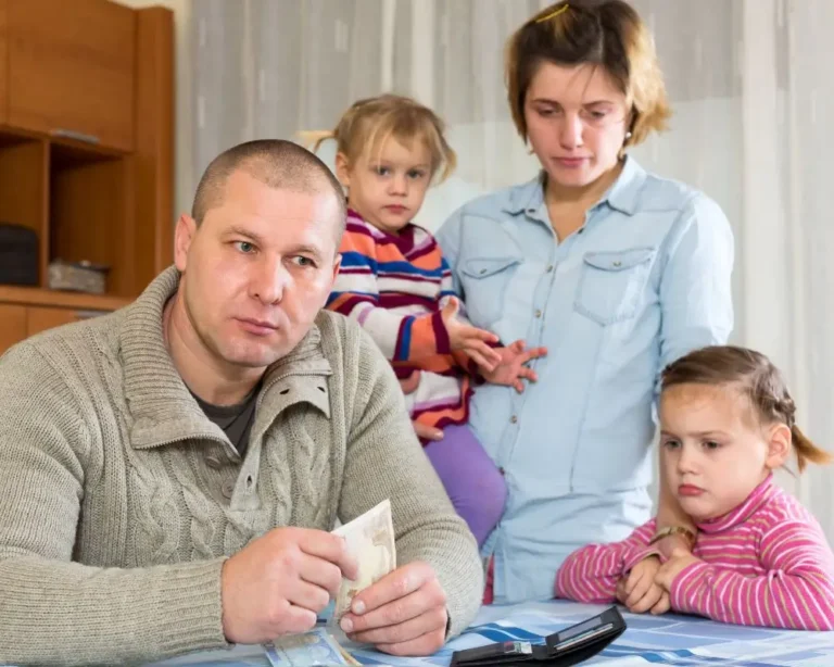 Zmartwiona rodzina przy stole – ojciec w szarym swetrze trzyma w dłoni banknot i patrzy w dal z poważną miną, obok niego siedzi smutna dziewczynka, a w tle stoją matka z drugą córką na rękach. Na stole leży cienki portfel i kilka banknotów, co symbolizuje trudną sytuację finansową i niepokój związany z brakiem alimentów.