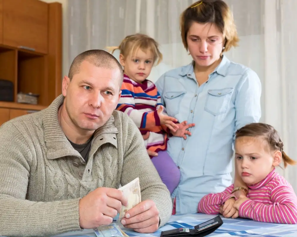 Co zrobić, gdy ojciec nie płaci alimentów? 1 Zmartwiona rodzina przy stole – ojciec w szarym swetrze trzyma w dłoni banknot i patrzy w dal z poważną miną, obok niego siedzi smutna dziewczynka, a w tle stoją matka z drugą córką na rękach. Na stole leży cienki portfel i kilka banknotów, co symbolizuje trudną sytuację finansową i niepokój związany z brakiem alimentów.