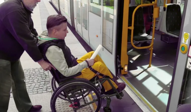 Czy opiekun osoby niepełnosprawnej płaci za bilet na przejazdy? 4 Mężczyzna pomaga osobie na wózku wjechać do niskopodłogowego tramwaju; otwarte drzwi, krawędź peronu i niewielki próg pokazują codzienne bariery, a gest wsparcia budzi poczucie bezpieczeństwa i ulgi. Scena z miejskiego przystanku ilustruje praktyczny kontekst pytania: czy opiekun osoby niepełnosprawnej płaci za bilet, oraz doświadczenie wspólnej podróży – skupienie, uważność, współpraca.