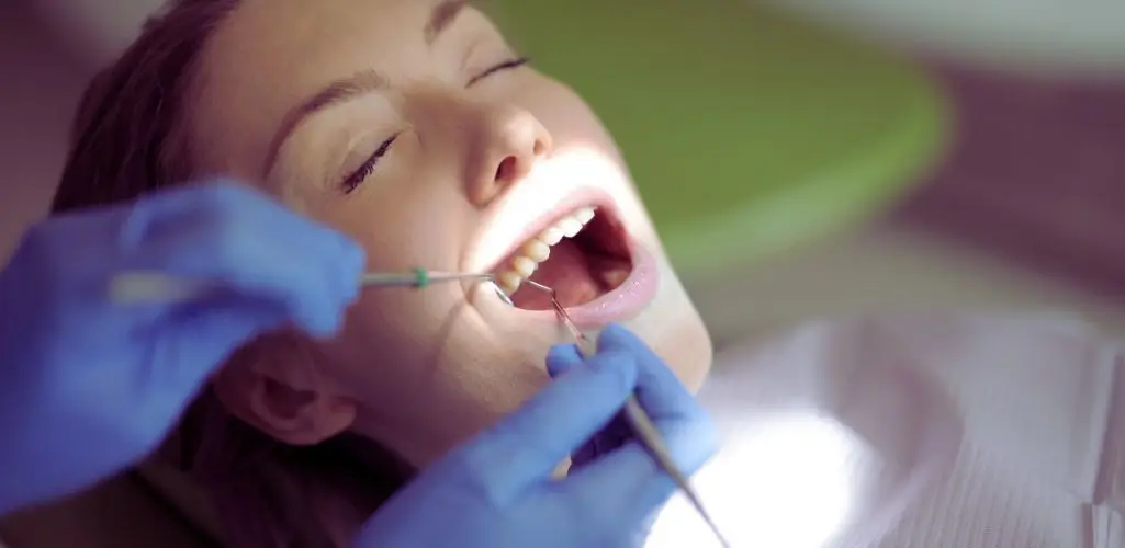 Kiedy NFZ refunduje leczenie zębów w narkozie dla niepełnosprawnych? 1 Dorosła pacjentka z zamkniętymi oczami leży na fotelu stomatologicznym, a dentysta w niebieskich rękawiczkach delikatnie bada jej zęby w jasnym świetle lampy; atmosfera spokoju i ulgi jak w sytuacjach, kiedy NFZ refunduje leczenie zębów w narkozie dla niepełnosprawnych.