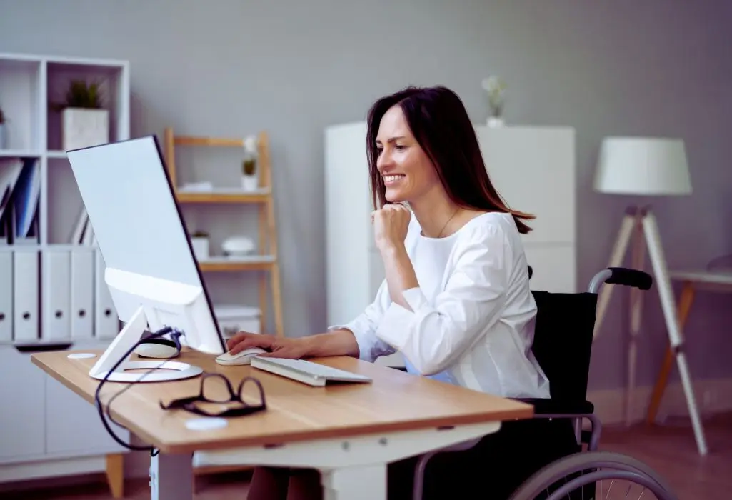 Uśmiechnięta kobieta na wózku przy komputerze, przygotowująca się do wyjścia i zastanawiająca się, co zabrać na rozmowę kwalifikacyjną.