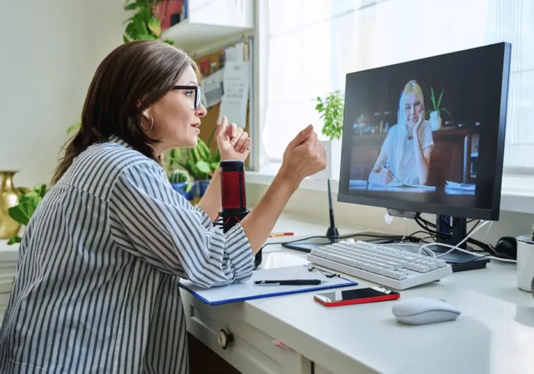 Kobieta w okularach rozmawia online z drugą kobietą widoczną na ekranie komputera. Siedzi przy biurku z mikrofonem i notatkami. Zdjęcie symbolizuje rekrutację i współpracę zdalną, które umożliwiają włączenie osób z niepełnosprawnościami w życie zawodowe.