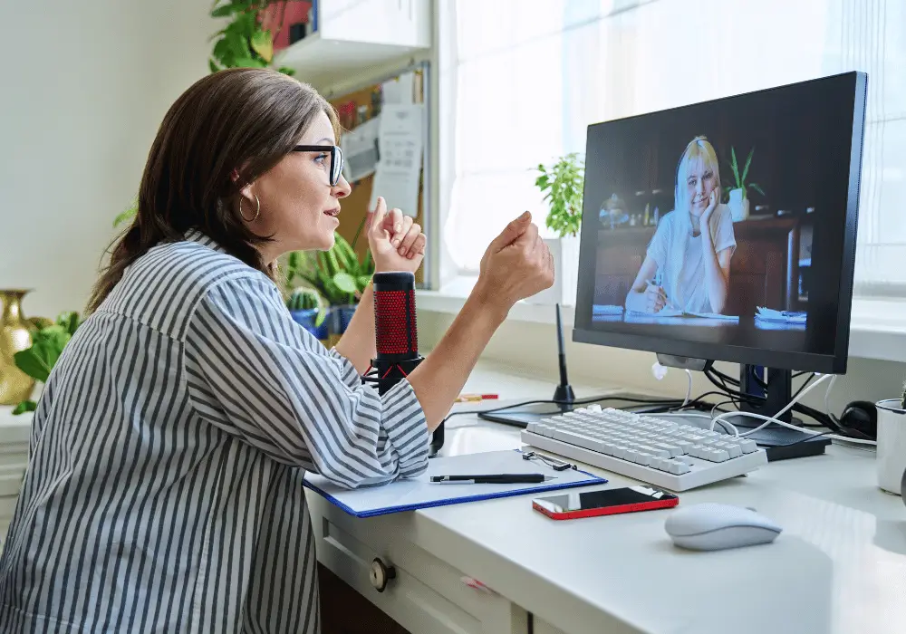 7 działań online, które przyciągną kandydatów z orzeczeniem o niepełnosprawności 1 Kobieta w okularach rozmawia online z drugą kobietą widoczną na ekranie komputera. Siedzi przy biurku z mikrofonem i notatkami. Zdjęcie symbolizuje rekrutację i współpracę zdalną, które umożliwiają włączenie osób z niepełnosprawnościami w życie zawodowe.