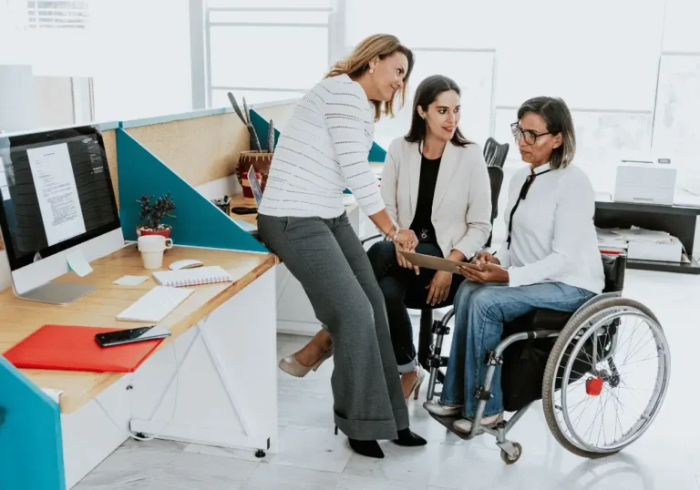 Trzy kobiety podczas rozmowy w biurze, jedna z nich siedzi na wózku inwalidzkim. Wspólnie przeglądają dokument na tablecie, uśmiechają się i współpracują przy biurku z komputerem. Zdjęcie ilustruje ideę integracji i równego udziału wszystkich członków zespołu w pracy.