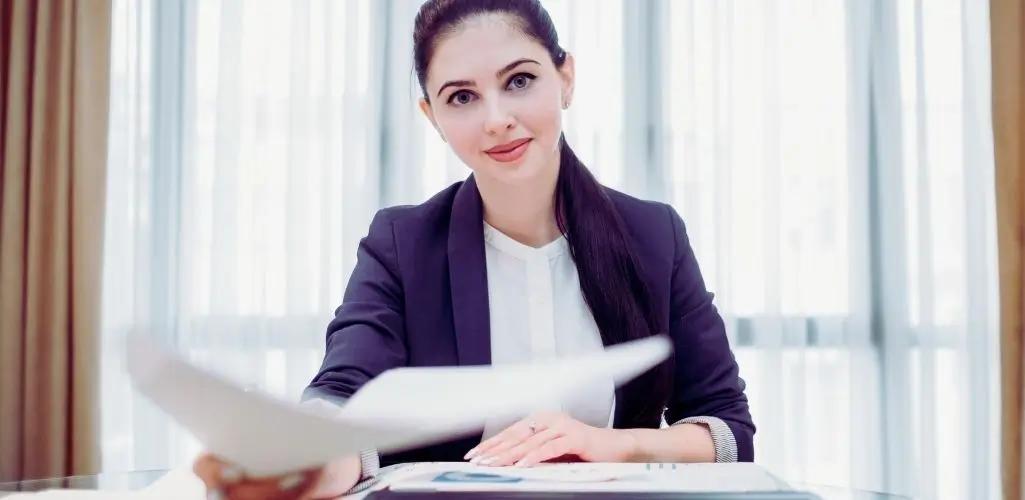 Kobieta w eleganckim stroju biurowym podaje dokument, symbolicznie pokazując, jak napisać podanie o pracę.