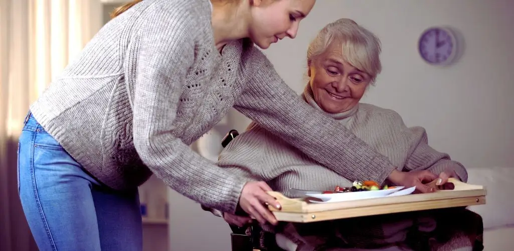 Opieka nad osobą niepełnosprawną – opiekunka podaje posiłek starszej kobiecie siedzącej na wózku inwalidzkim.